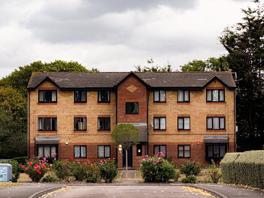 A three-story brick residential building with a dark tiled roof, featuring multiple windows with black frames and white curtains. In front of the building, there is a well-maintained garden with vibra