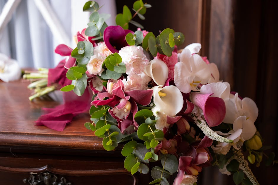 A floral bouquet arrangement featuring pink and white calla lilies, pink and white carnations, deep burgundy calla lilies, and sprigs of green eucalyptus with round leaves, all set on a polished woode