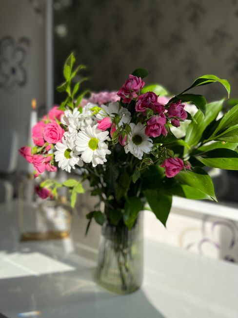 A fresh flower bouquet arranged in a clear glass vase placed on a white surface, featuring a combination of pink roses, white daisies with yellow centers, and small pink and purple blooms, complemente