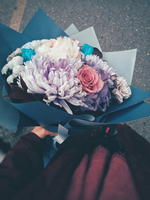 A beautifully arranged bouquet of fresh flowers held by a person outdoors, featuring a large white and purple chrysanthemum, a pink rose, and smaller white and blue flowers, all wrapped in elegant blu
