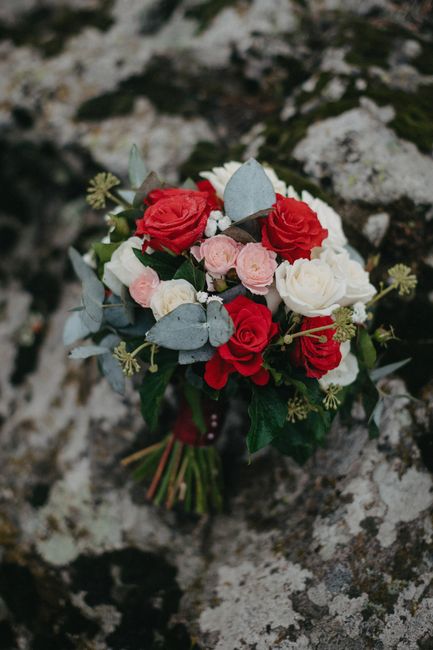 A bouquet of fresh flowers featuring red and white roses, complemented by soft pink blooms and sprigs of silvery eucalyptus leaves, arranged in a rounded, lush style. The bouquet is wrapped with a dar