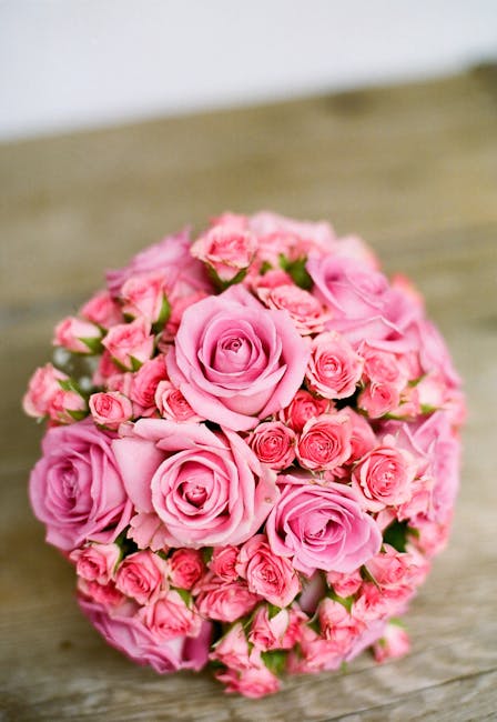 A round bouquet of fresh pink roses and smaller pink bud flowers arranged closely together, with smooth, velvety petals and green stems visible at the base. The bouquet is placed on a rustic wooden su